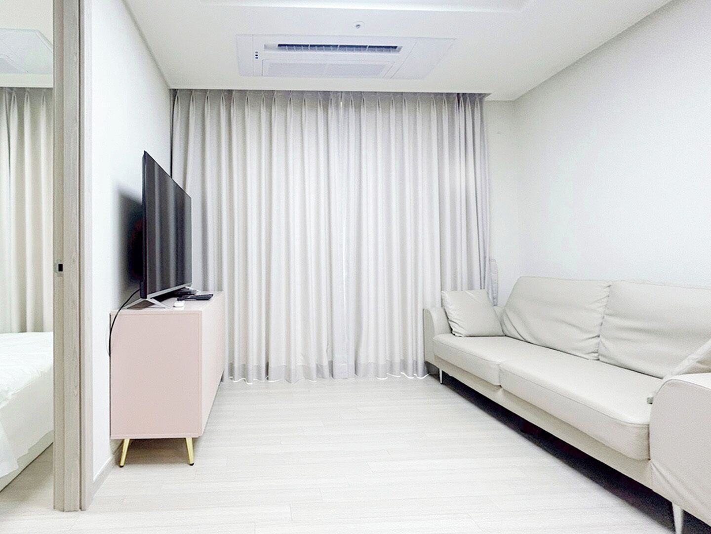 Una habitación de hotel moderna con un sofá blanco, un televisor de pantalla plana sobre un mueble rosa y un ventanal con cortinas blancas.