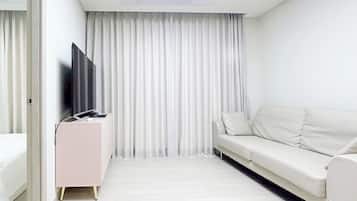 Una habitación de hotel moderna con un sofá blanco, un televisor de pantalla plana sobre un mueble rosa y un ventanal con cortinas blancas.
