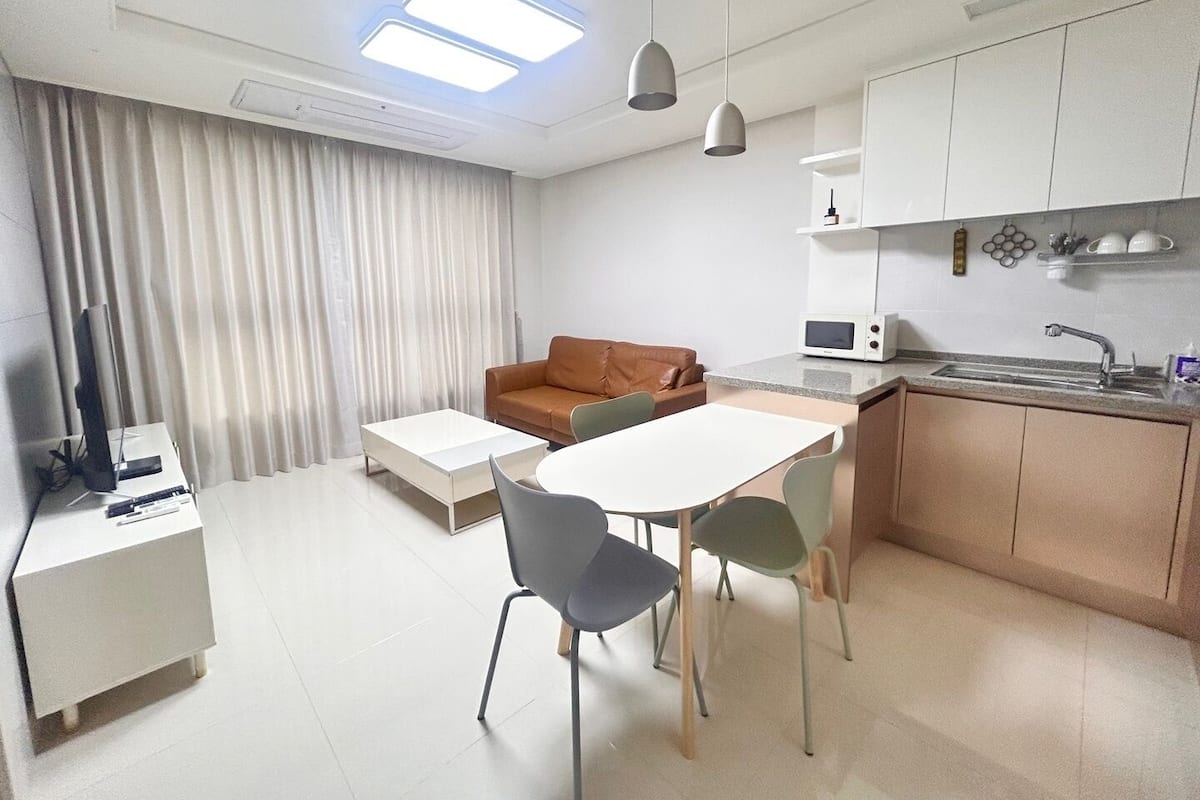 Ein modernes, kompakter Wohnraum mit Küchenzeile, einer Essecke mit Tisch und Stühlen, einer Couch und einem Fernseher auf einem Schrank.