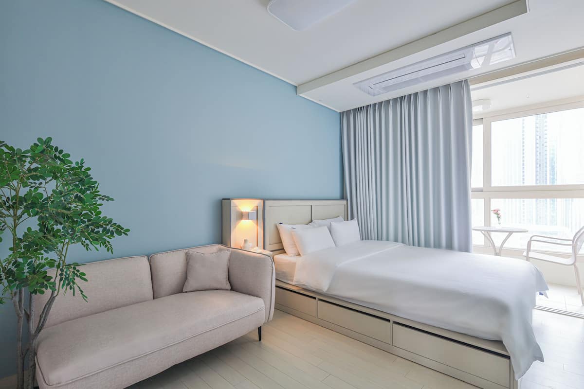 Ein Hotelzimmer mit Bett, Sofa, Nachttisch, Lampe, einem Fenster mit Vorhängen und einer Zimmerpflanze.