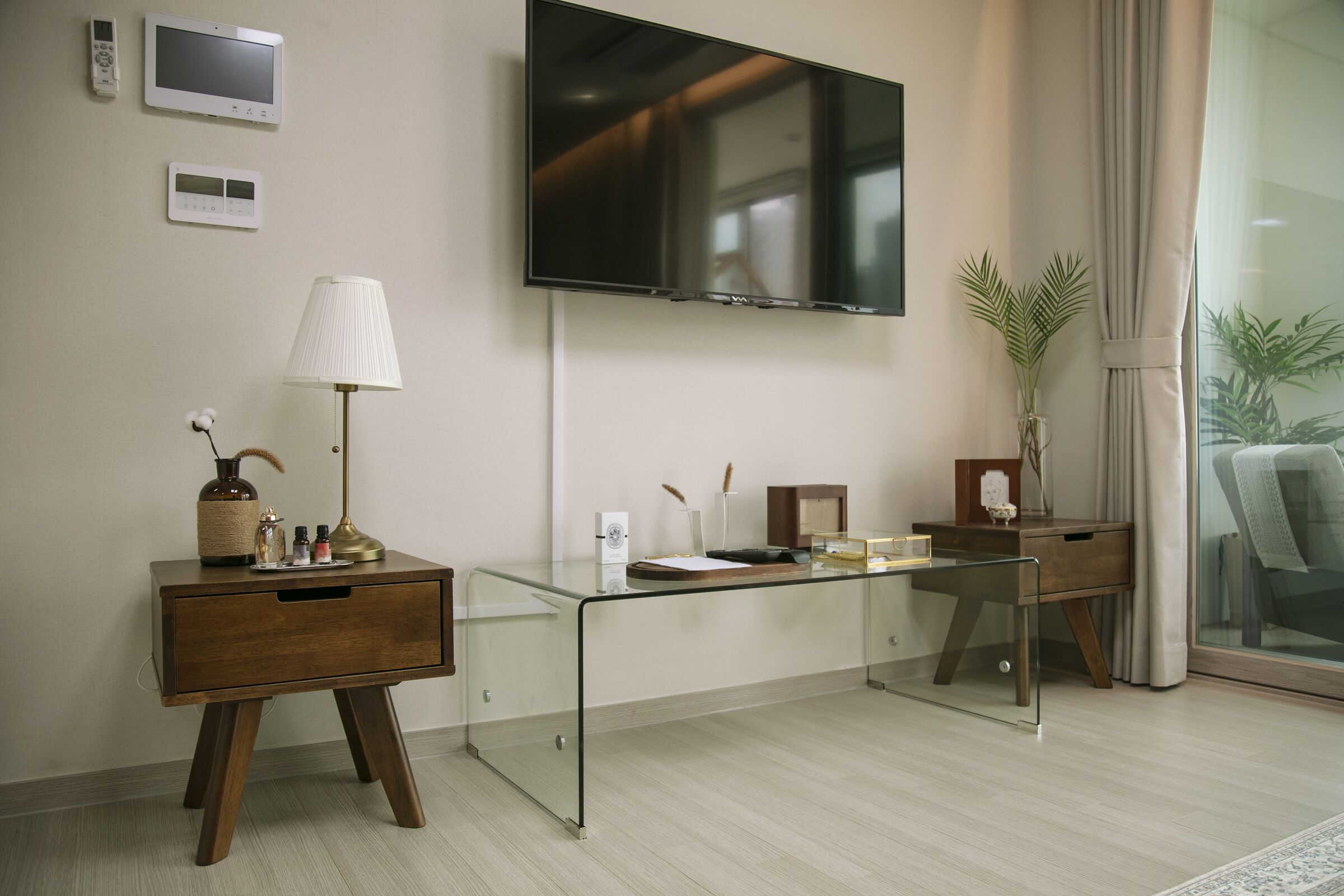 A modern hotel room with a glass desk, a wooden nightstand, a flat-screen TV, and a large window with curtains.