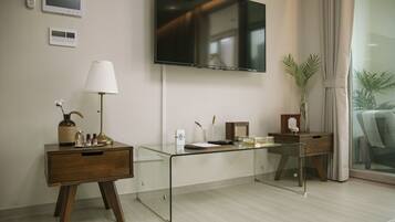 A modern hotel room with a glass desk, a wooden nightstand, a flat-screen TV, and a large window with curtains.