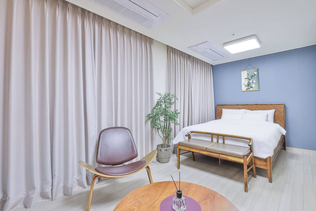 Ein Hotelzimmer mit Bett, Sessel, kleinem Tisch, Blumentopf und Vorhängen.