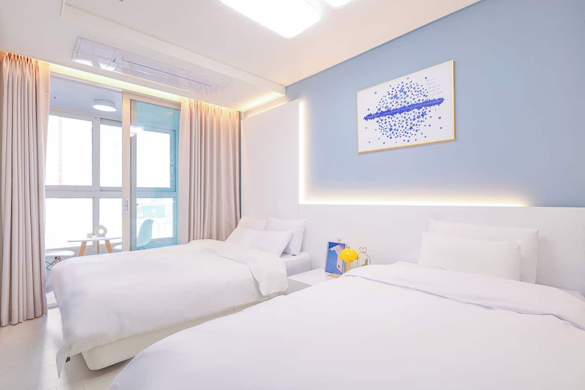 Ein Hotelzimmer mit zwei Betten, einem großen Fenster mit Vorhängen, einem Wandbild und einem kleinen Tisch mit einem Stuhl.