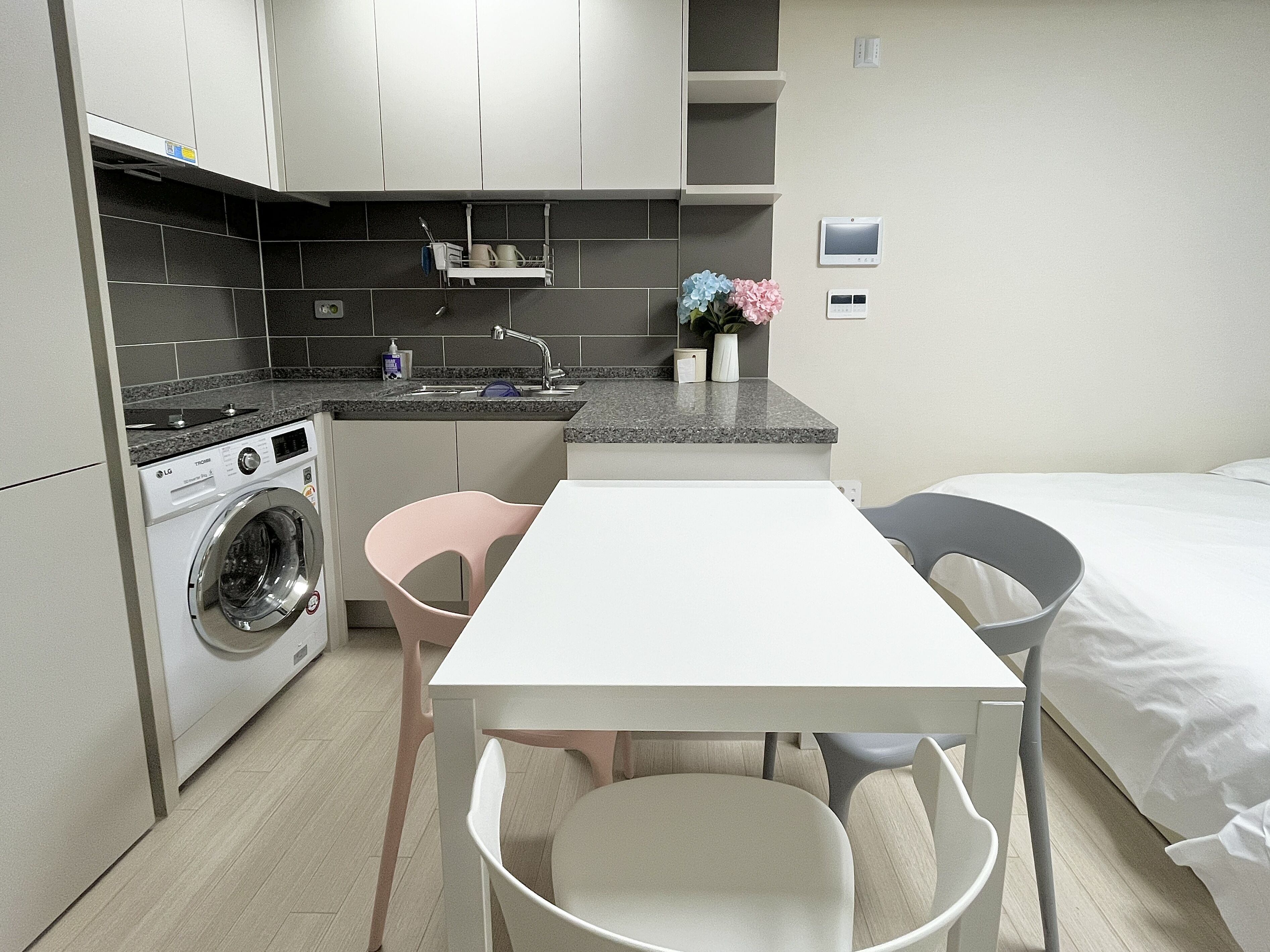 Une cuisine compacte équipée d’un lave-linge, d’un évier et d’un petit plan de travail. On y trouve une table avec des chaises et un lit avec du linge de lit blanc.
