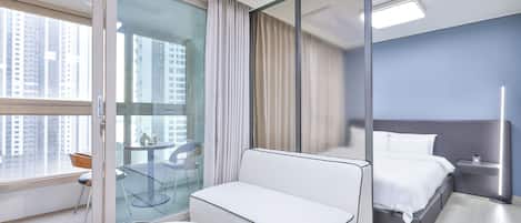 A modern hotel room with a bed, a sofa, a glass partition, a small table, and a chair.