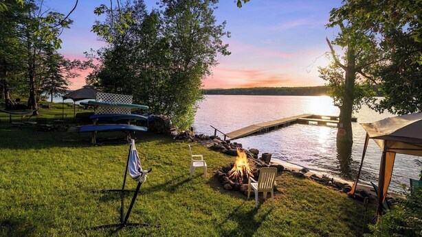 A lakeside camping area with a fire pit, picnic tables, and a dock extending into the water.