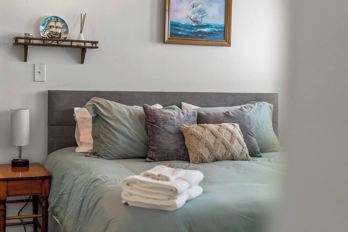 A neatly made bed with a grey headboard, multiple pillows, and a folded towel on the side. A small wooden nightstand with a lamp and a decorative plate is beside the bed.