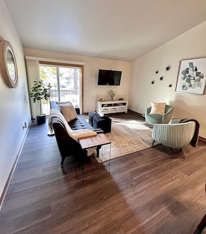 Un salon moderne avec un canapé, des fauteuils, une table basse, un téléviseur et une grande fenêtre.