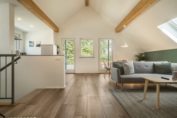 A modern interior with a loft-style ceiling, a grey sofa, a wooden dining table, and a kitchen area.