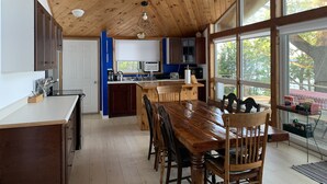 A spacious kitchen with wooden ceiling, a dining area with a large wooden table, and a view of the outdoors through large windows. - Water Access Only Cottage on Georgian Bay (Tay)