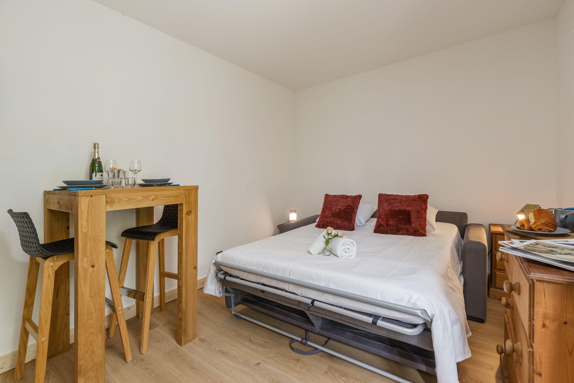 A compact bedroom with a bed, a desk with chairs, a small table with a bottle and glasses, and a dresser with a lamp and a plate of food.