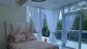 A bedroom with a bed, a large window with curtains, and a view of greenery outside. - New lovely house at walking distance from beautiful Sosua beach.  (Sosúa)