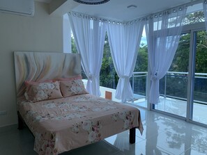 A bedroom with a bed, a large window with curtains, and a view of greenery outside.