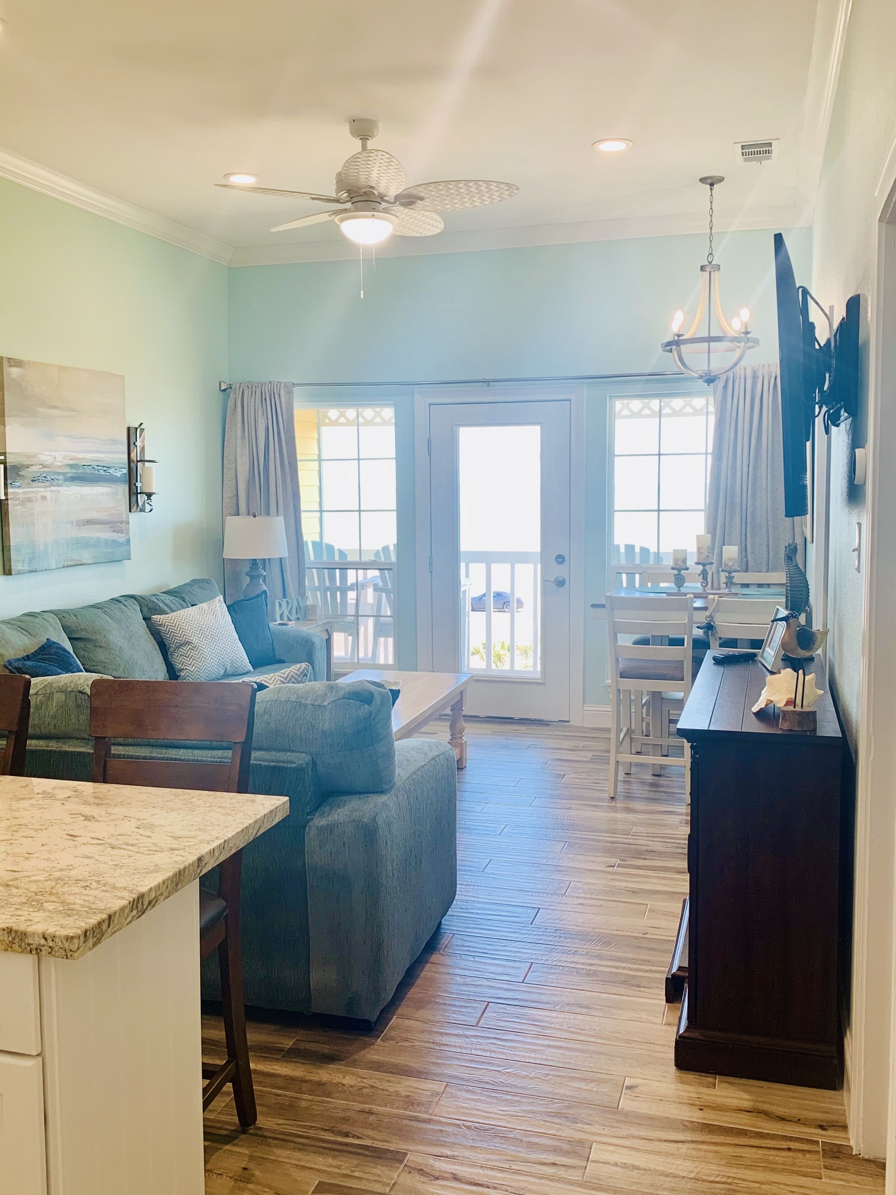 A spacious living area with a sofa, dining table, and chairs. There is a balcony with a view, and a ceiling fan.