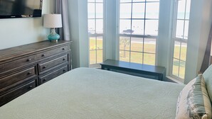Una camera da letto con un letto, una cassettiera, una lampada e vista sulla spiaggia attraverso ampie finestre.