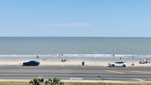 Una spiaggia con una strada in primo piano, alcune auto e persone che si godono il mare.