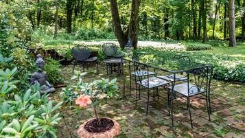 A garden patio with metal chairs and tables, surrounded by lush greenery and trees.