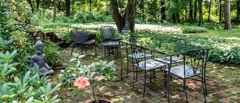 A garden patio with metal chairs and tables, surrounded by lush greenery and trees.