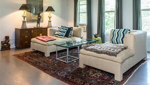 A living room with a sofa, ottoman, glass coffee table, and a patterned rug. There are two lamps, a decorative statue, and a framed artwork on the wall.