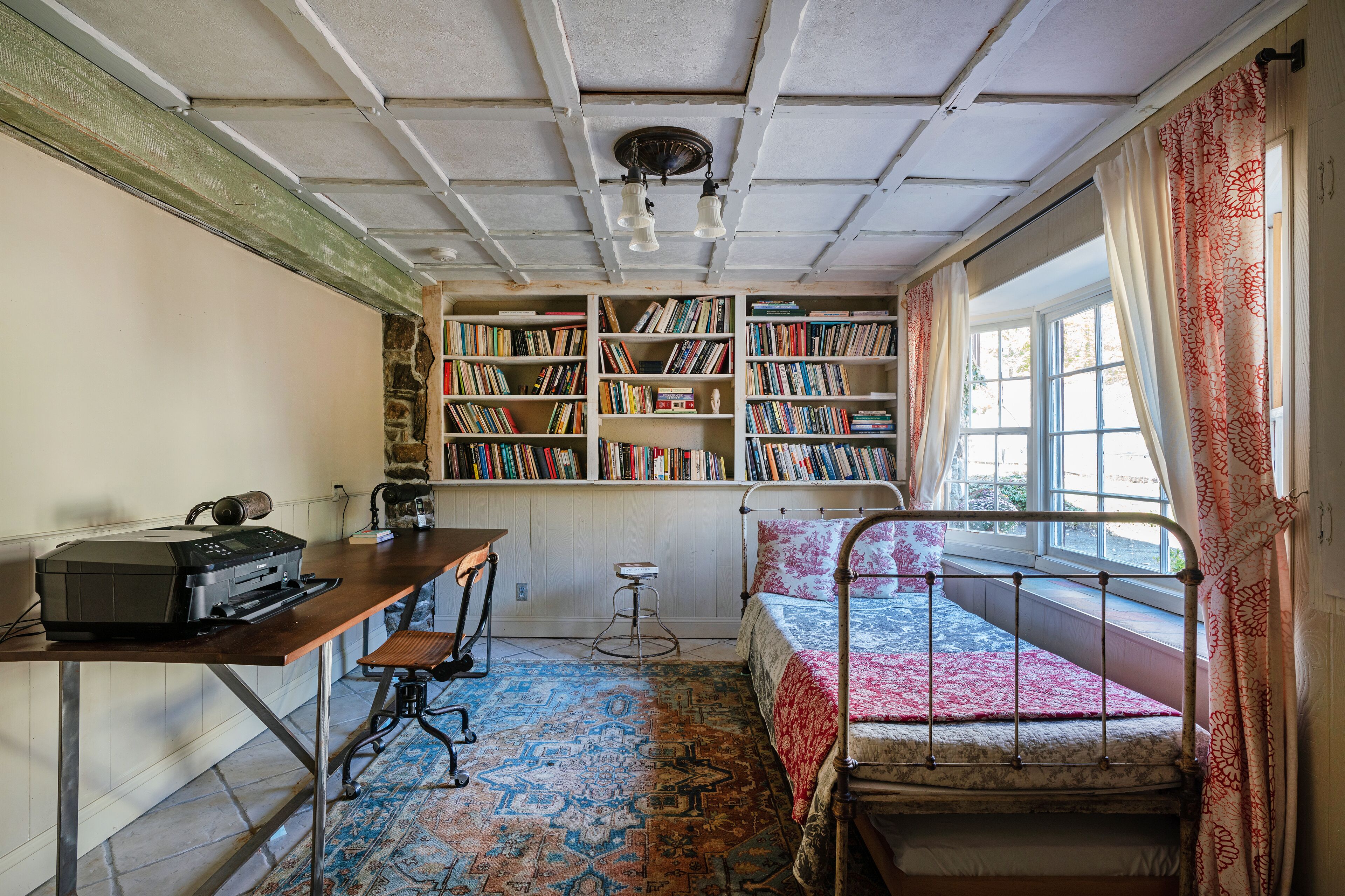 Een kamer met een bed, een bureau met een printer en boekenkasten vol boeken.