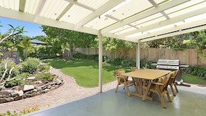 Une terrasse couverte avec une table et des chaises, un barbecue et un chemin de pierres menant à une cour gazonnée.