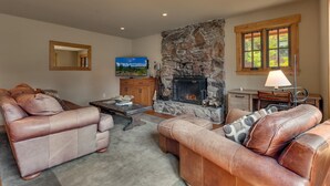 Una sala de estar con sofás de cuero, una chimenea de piedra, un televisor y un ventanal con vistas a árboles.