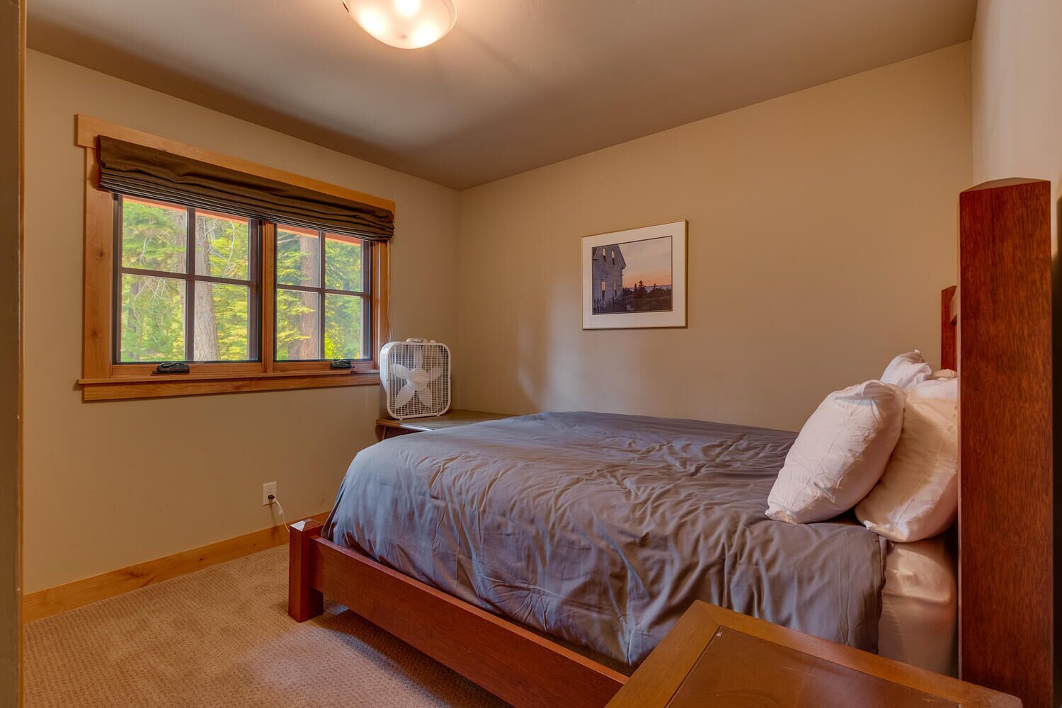 Un dormitorio con cama de madera, un ventilador de pie, una ventana con vistas a árboles y un cuadro enmarcado en la pared.