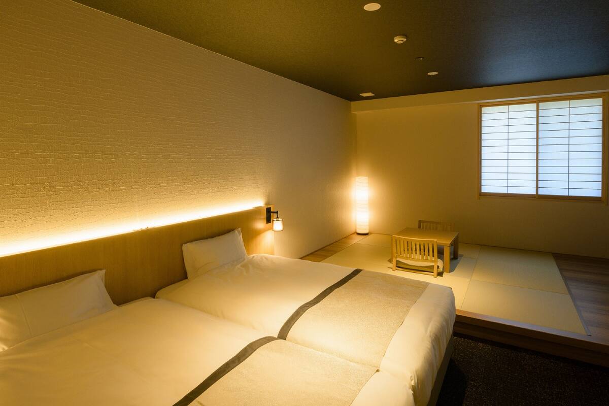 Une chambre d’hôtel avec un grand lit, des tables de chevet, un petit coin salon et une fenêtre avec des stores.