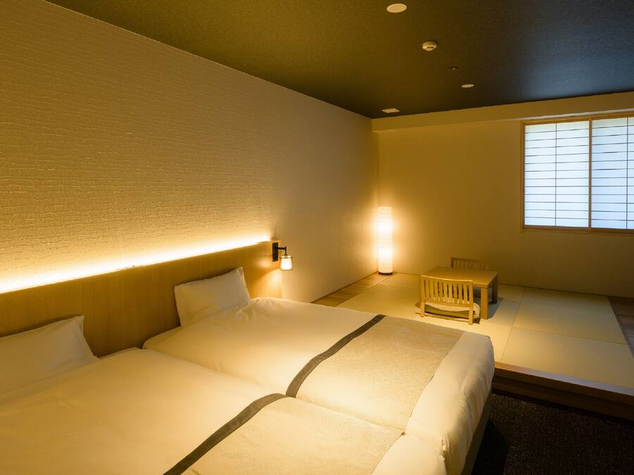 Une chambre d’hôtel avec un grand lit, des tables de chevet, un petit coin salon et une fenêtre avec des stores.
