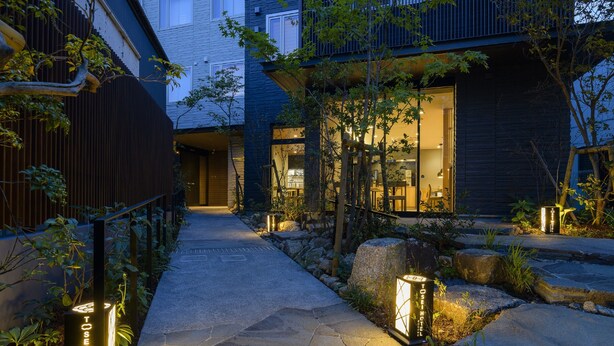 Un sendero de piedra conduce a un edificio moderno con fachada oscura y puertas de cristal, rodeado de follaje y detalles de piedra.