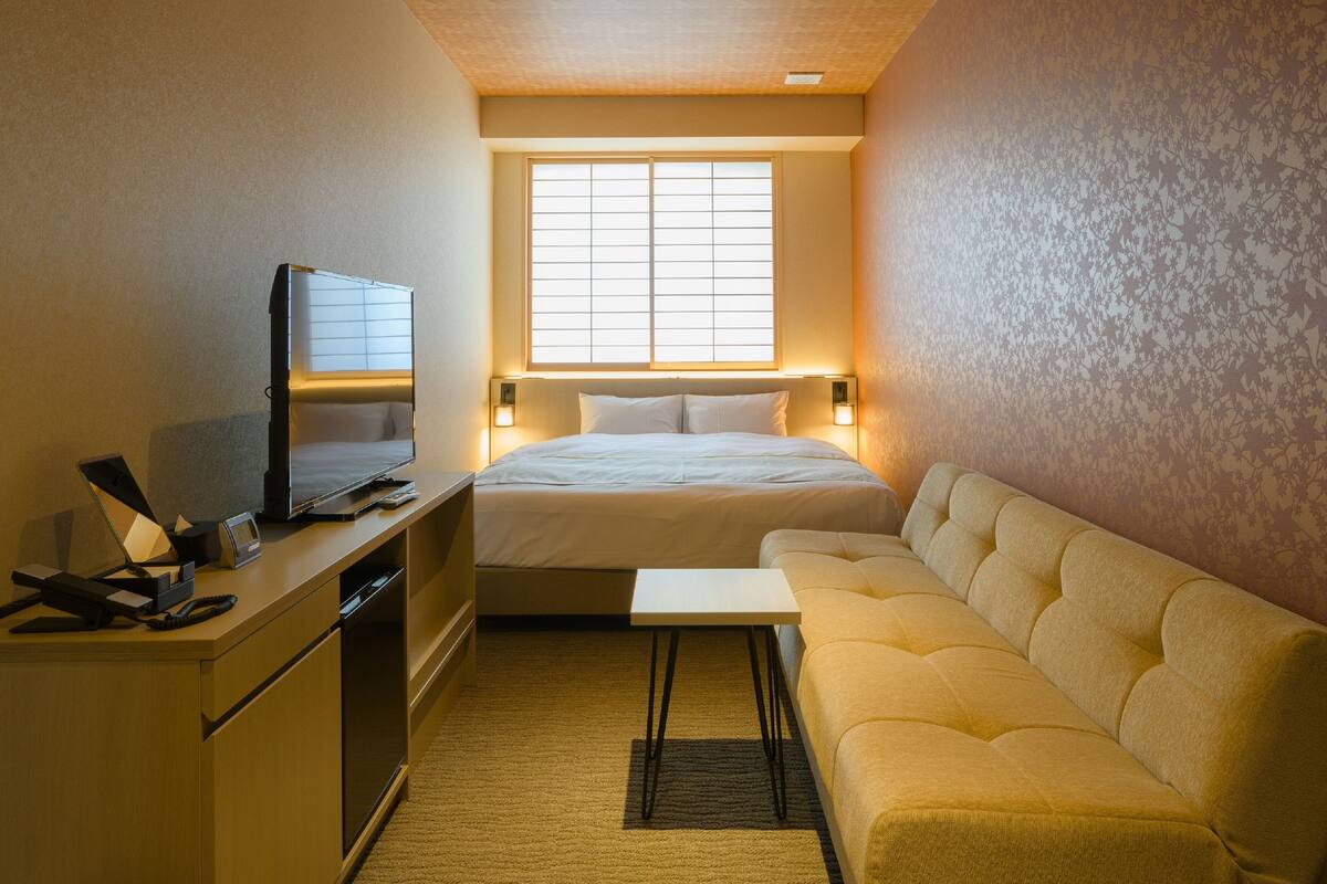 Une chambre d’hôtel compacte comprenant un lit, un canapé, un bureau avec une télévision et une fenêtre laissant entrer la lumière naturelle.