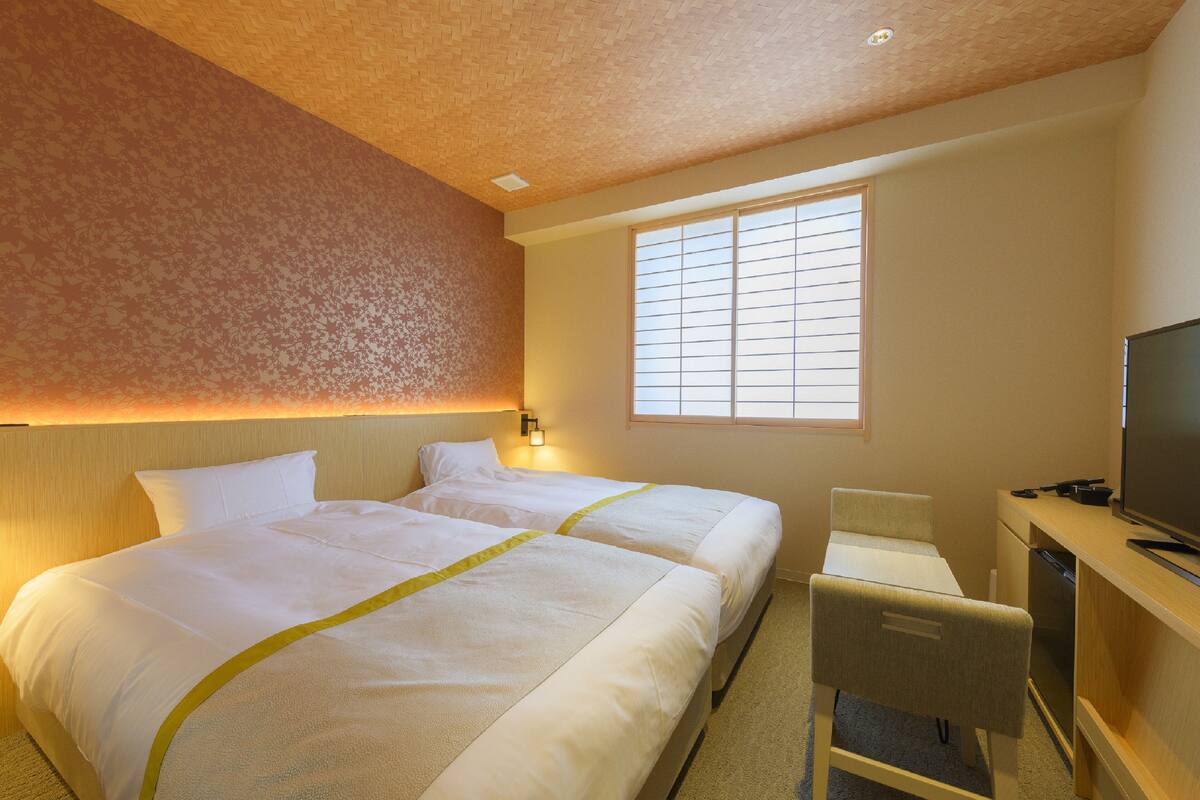Une chambre d’hôtel avec deux lits, un bureau avec une télévision et une fenêtre avec des stores.