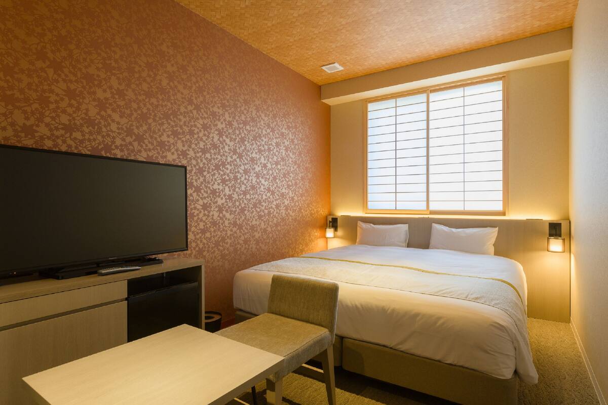 Une chambre d’hôtel équipée d’un lit, d’une télévision, d’un bureau et d’une fenêtre avec des stores.