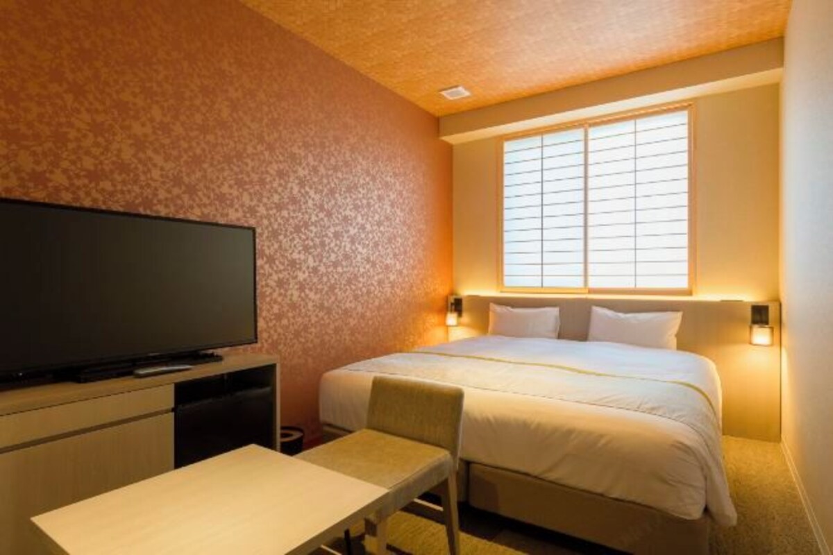 A hotel room with a bed, a television, a desk, and a window with blinds.