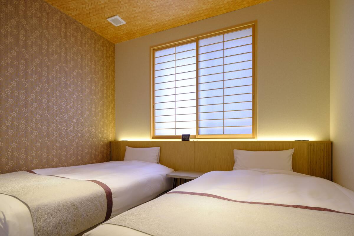 Une chambre d’hôtel avec deux lits, une grande fenêtre avec des stores et un papier peint à motifs sur un des murs.