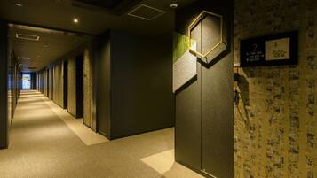 A hotel corridor with a carpeted floor, a wall-mounted sign with room numbers, and a patterned wallpaper on the right wall.