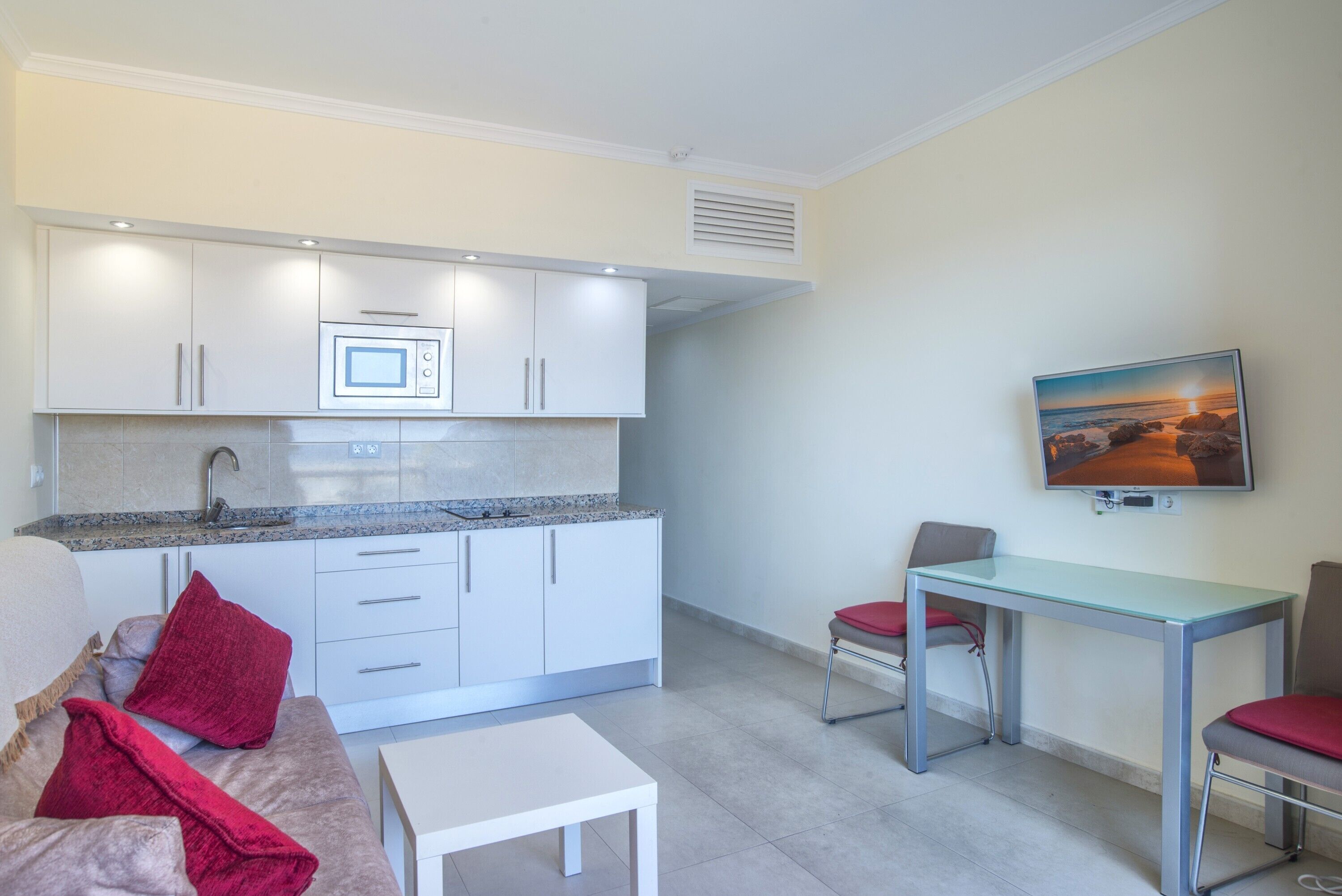 Un espacio habitable compacto con kitchenette, comedor, sofá con cojines rojos y televisor montado en la pared.