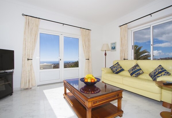 A living room with a sofa, a coffee table, a TV, and a view of the ocean.