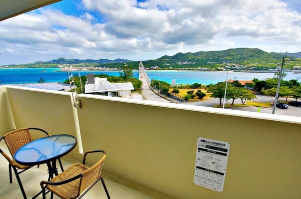 A balcony with wicker chairs and a small table, overlooking a coastal view with mountains and a bridge.