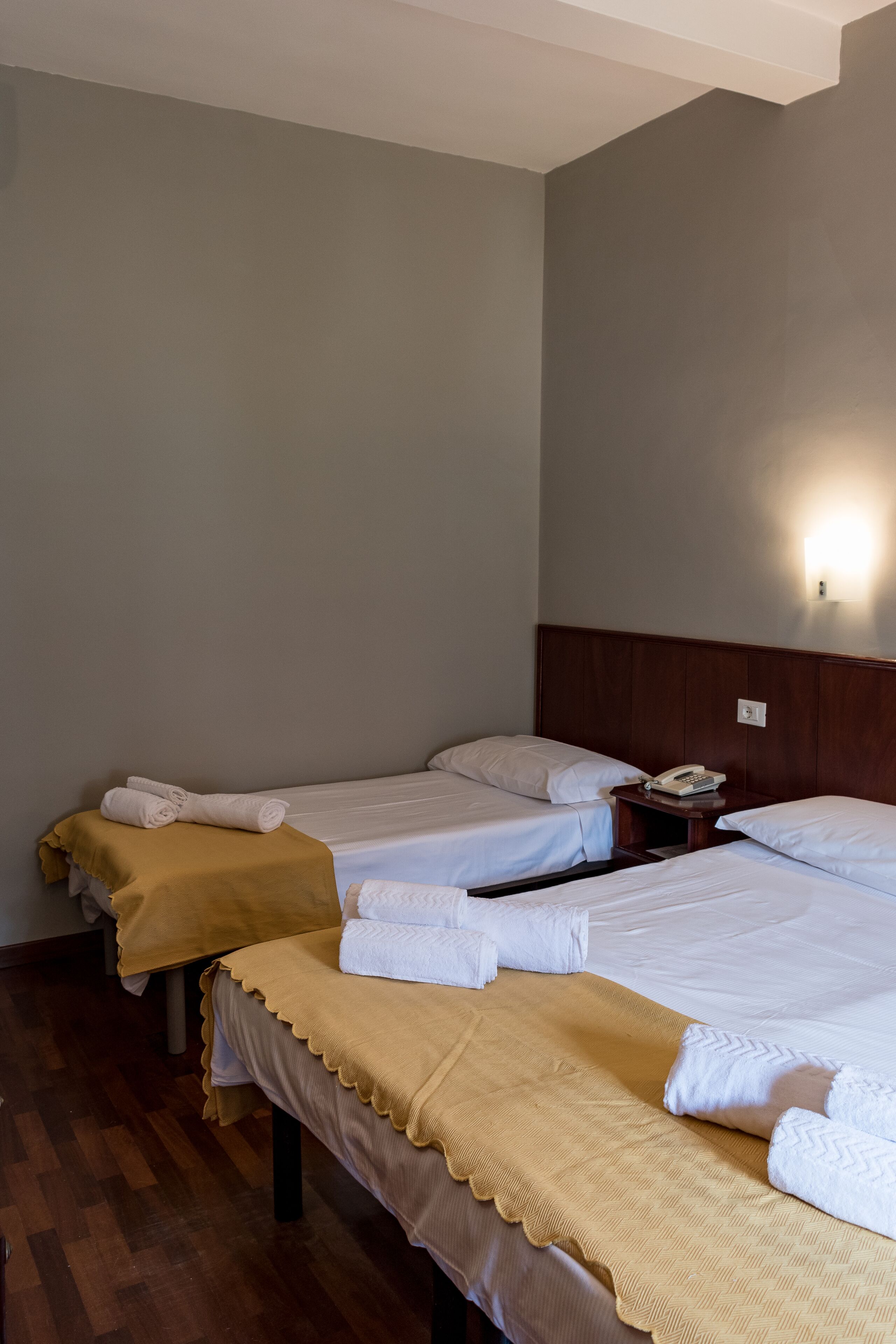 A hotel room with two beds, each with white linens and a brown throw blanket. There is a wooden nightstand with a telephone and a wall-mounted light fixture.