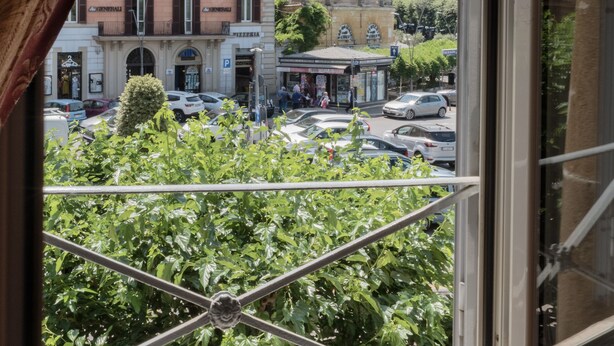 A window view of a building with a balcony, a street with cars, and a hill in the background. - Hotel Bellavista (Frascati)
