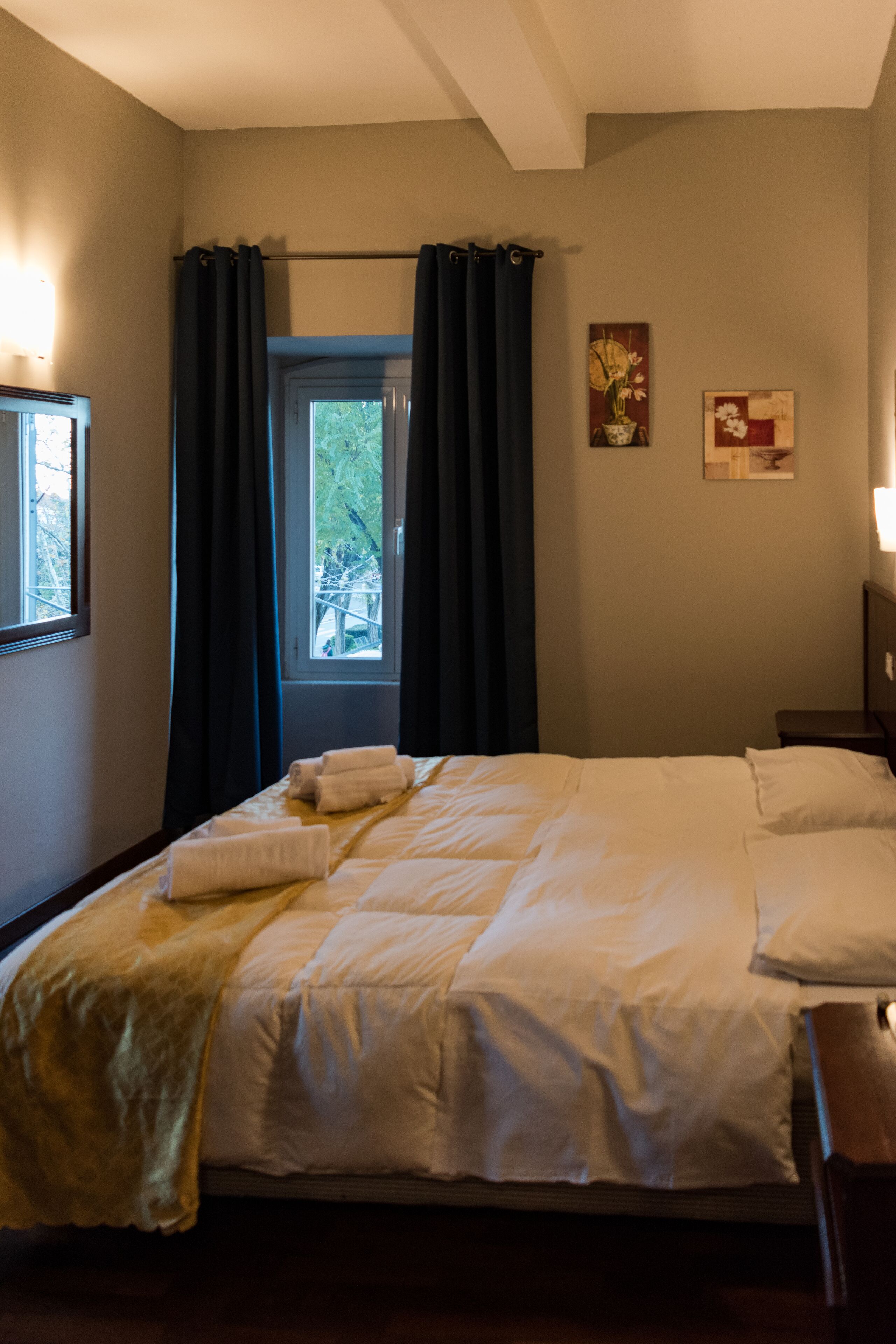A neatly made bed with white linens and a yellow throw blanket, a window with dark curtains, and two wall-mounted paintings.