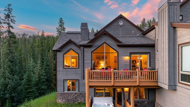 A two-story lodge with a balcony, stone foundation, and a hot tub.
