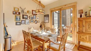 A dining area with a wooden table set for four, surrounded by chairs. There are framed pictures on the wall, a chandelier, and a wooden cabinet with a decorative top.