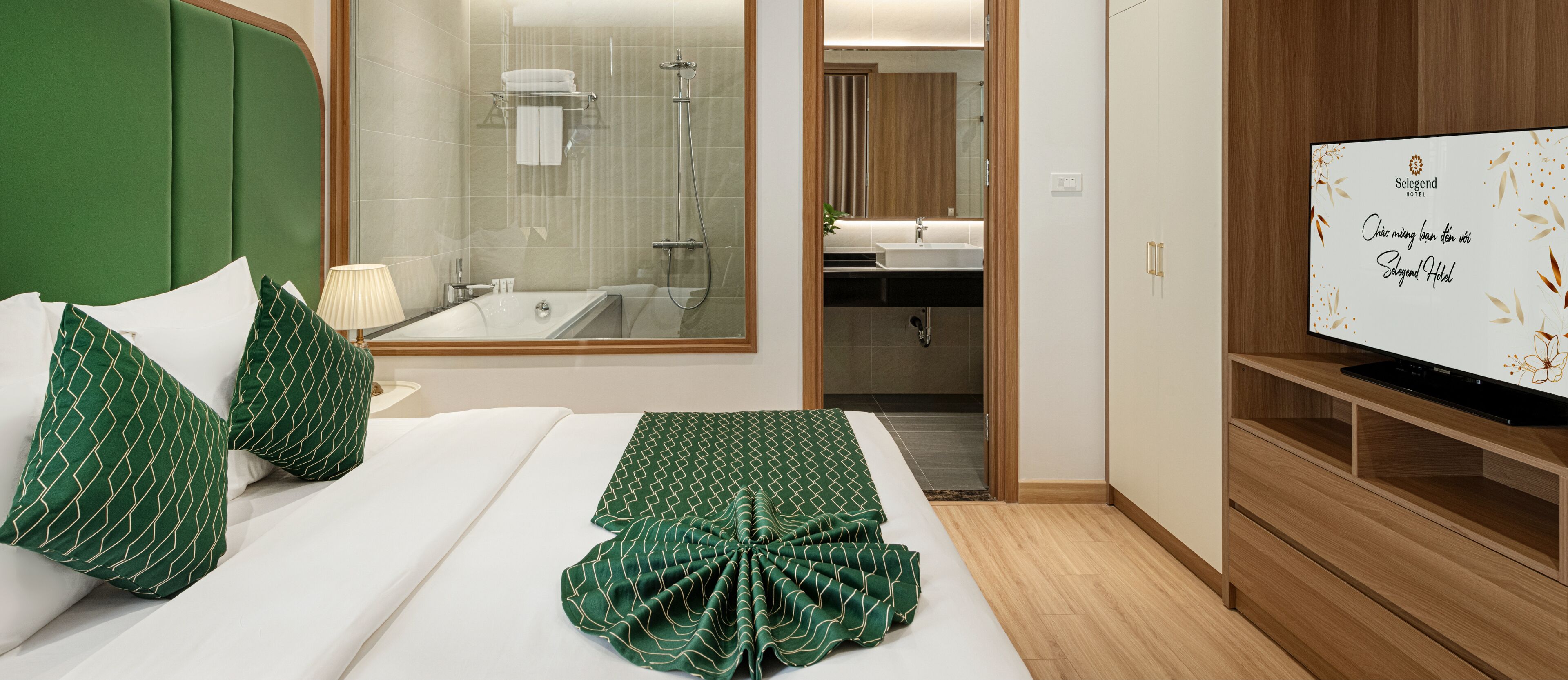 A hotel room with a bed, green pillows, a TV, a bathroom with a shower, and a wooden cabinet.