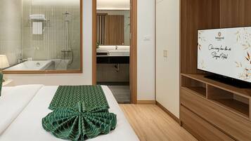 A hotel room with a bed, green pillows, a TV, a bathroom with a shower, and a wooden cabinet.