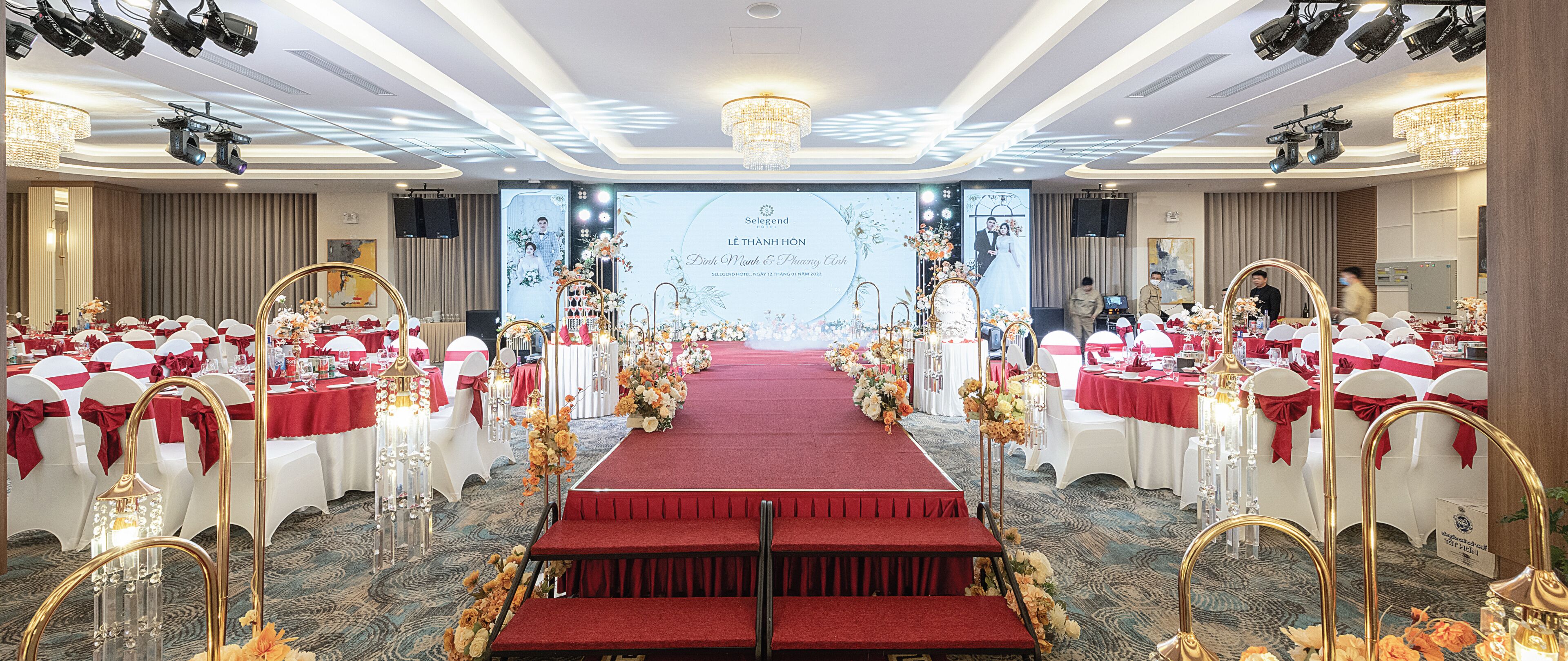 A hotel ballroom with a red carpet, white chairs with red sashes, and floral decorations.