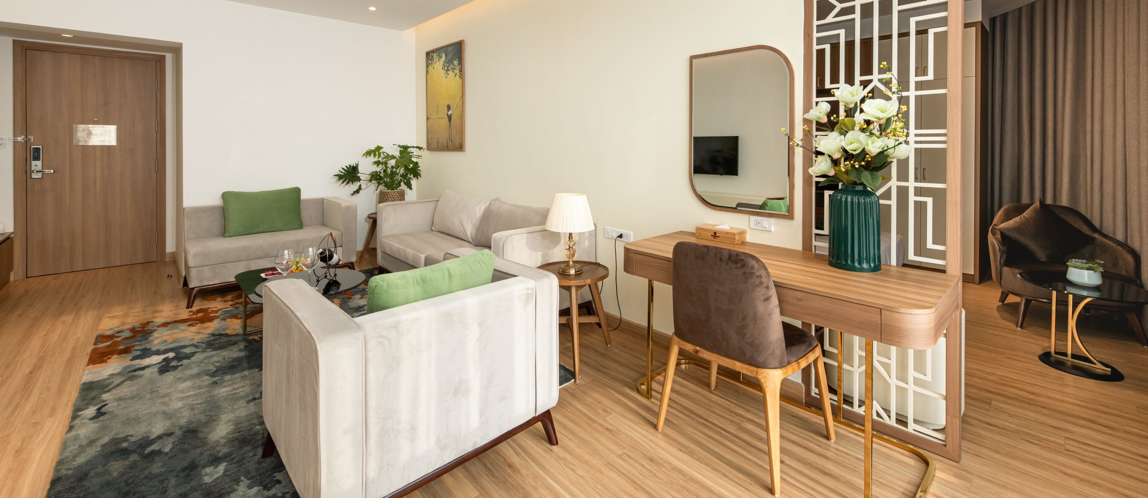 Ein modernes Hotelzimmer mit Sofa, Schreibtisch, Stuhl, einem kleinen Tisch mit Lampe, einer Vase mit Blumen und einem Spiegel.