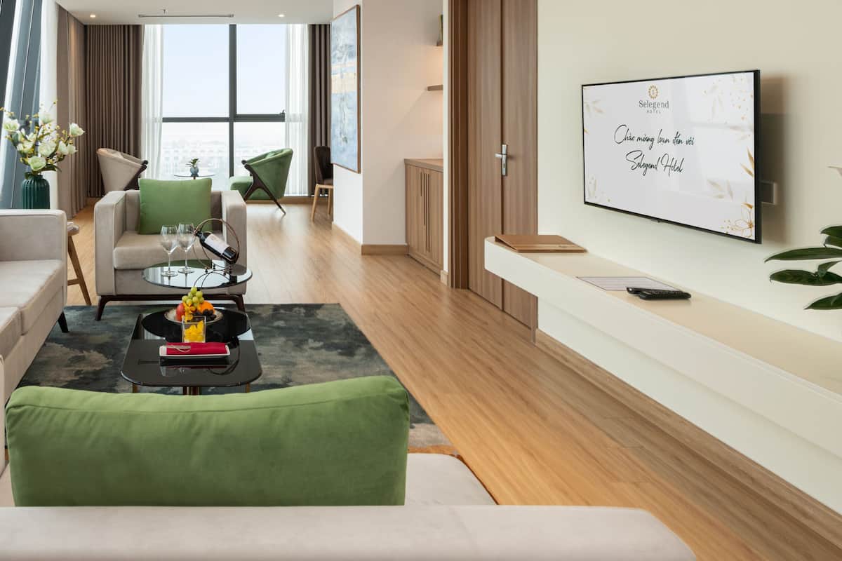 Un salon moderne avec un canapé, un fauteuil et une table basse, une grande fenêtre donnant sur la ville et un téléviseur fixé au mur.